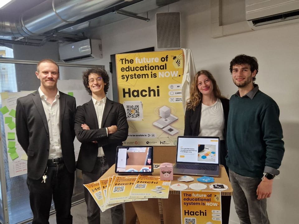 The four members of the Hachi team standing beside their stand, with the Hachi poster, printed learning cards, a tablet and a laptop.